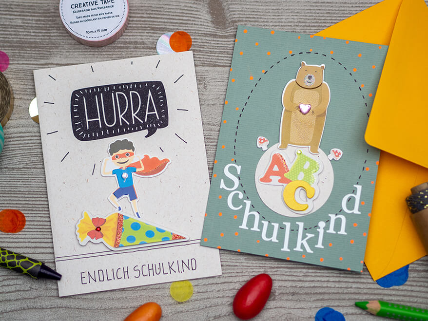 Auf einem hellgrauen Holzuntergrund liegen eine wasserblaue Grußkarte auf die ein brauner Bär, die Buchstaben ABC und das Wort Schulkind mit Aufklebern der Firma Rössler geklebt ist. Daneben liegt eine weiße Karte, mit einem gestempelten "Hurra", einem Superhelden-Kind, das auf einer Schultüte steht und der Spruch "Endlich Schulkind" drauf. Drumherum liegen buntes Konfetti, eine Garnspule, Washitape und verschiedene Stifte verteilt.