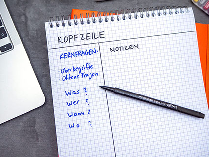 Auf einem dunklen Untergrund liegt ein orangefarbenes Notizbuch, darauf ein karierter Notizblock. Auf dem Block ist die Aufteilung für die Cornell Methode gezeichnet. Der Bereich mit den Kernfragen ist mit blauem Stift farblich hervorgehoben. Auf dem Block liegt ein Fasermaler der Marke Staedtler.