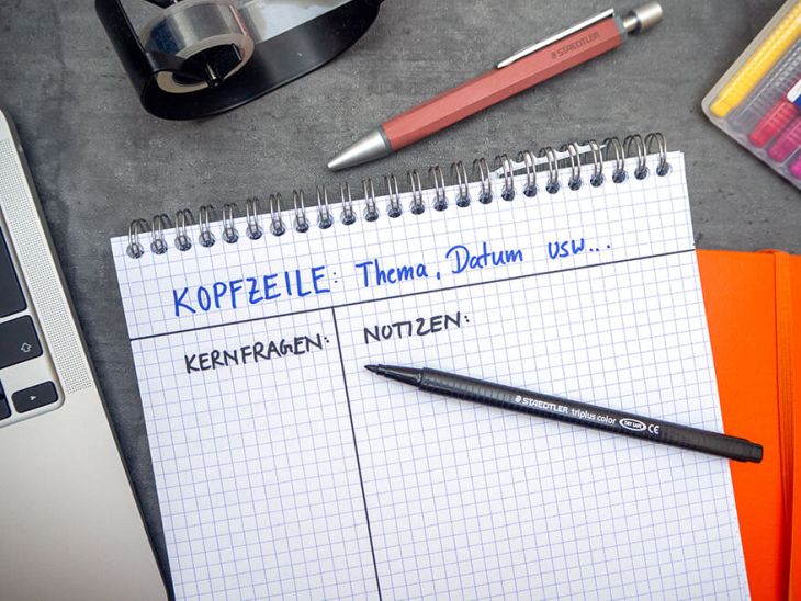 Auf einem dunklen Untergrund liegt ein orangefarbenes Notizbuch, darauf ein karierter Notizblock. Auf dem Block ist die Aufteilung für die Cornell Methode gezeichnet. Die Kopfzeile ist mit blauem Stift farblich hervorgehoben. Auf dem Block liegt ein Fasermaler der Marke Staedtler. Drumherum ist weiteres Büromaterial verteilt.