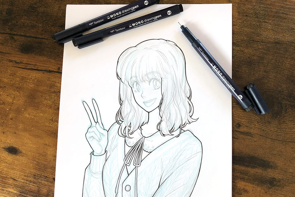 Auf einem braunen Holztisch liegt ein Blatt Papier. Darauf wurde die Skizze einer jungen Frau im Manga-Stil mit Finelinern von Tombow nachgezeichnet. Foto: Reyhan Yildirim