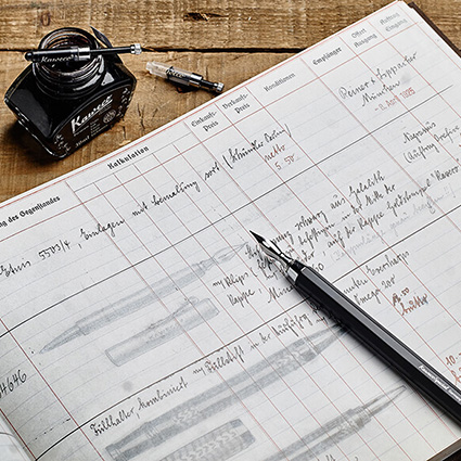 Auf einem rustikalen Holzuntergrund liegt ein aufgeschlagenes, beschriebenes Notizbuch. Darauf liegt ein schwarzer Kaweco Special DipPen. Oberhalb davon ist ein offenes, dunkles Tintenfass zu sehen.