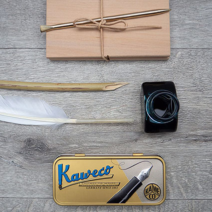 Auf einem rustikalen Holzuntergrund liegen von oben nach unten: eine römische Wachstafel mit einem Griffel aus Bronze, ein angespitztes Schilfrohr (Calamus), eine Gänsefeder, ein Tintenfass von Aratrum und eine Blechdose mit einem modernen Füllfederhalter von Kaweco. Foto: Johanna Rundel