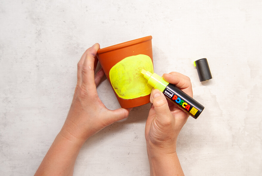 Jemand malt mit einem dicken, gelben Marker von Posca ein Oval auf einen Blumentopf aus Ton. Der Untergrund ist hell. Foto: Johanna Rundel