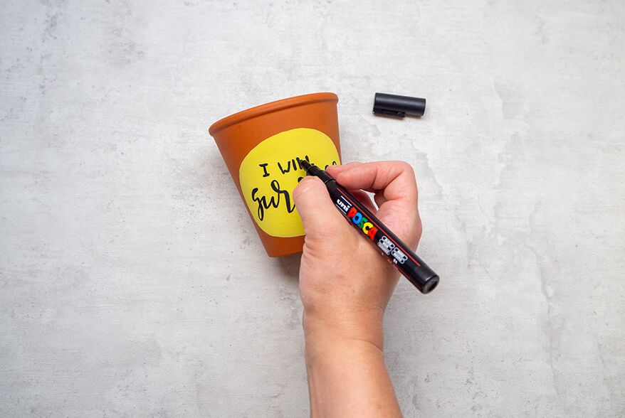 Jemand schreibt auf einen Blumentopf aus Ton mit einem schwarzen Marker von Posca den Satz „I will survive“. Der Untergrund ist hell. Foto: Johanna Rundel