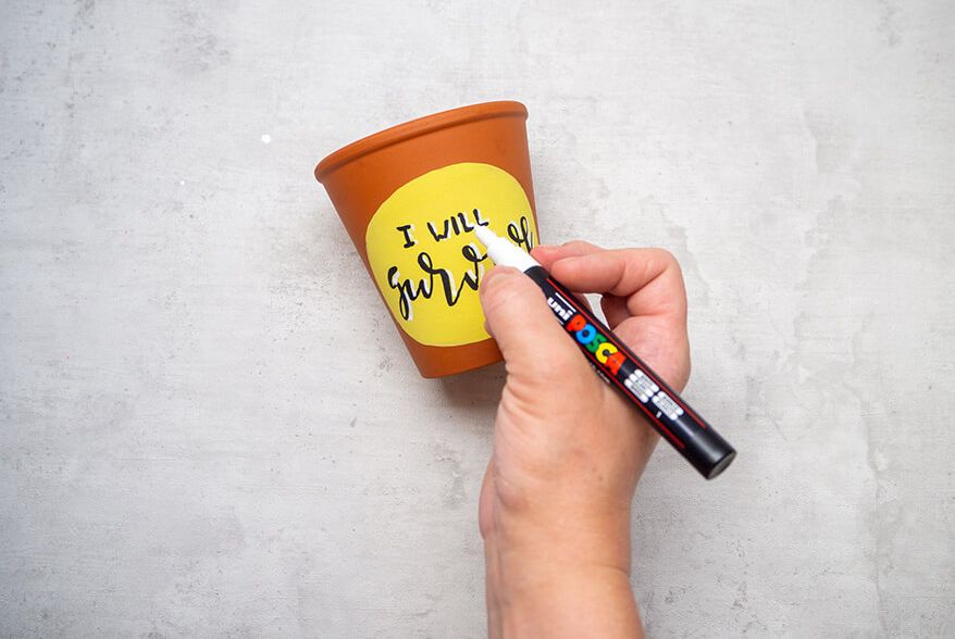 Jemand setzt mit einem weißen Marker von Posca Akzente auf das Lettering „I will survive“, das auf einem gelben Oval auf einem Blumentopf aus Ton geschrieben wurde. Der Untergrund ist hell. Foto: Johanna Rundel