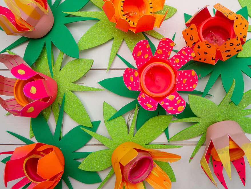 blumengirlande-draufsicht.jpg Auf einem hellen Untergrund sieht man von oben eine Lichterkette aus upcycelten Plastikflaschen in Blumenform liegen. Die Farben der Blüten sind Orange, Pink, Rosa und Gelb. Unter den Blüten sind grüne Blätter aus Moosgummi. Die Glühbirnchen der Lichterkette leuchten. Foto: Johanna Rundel