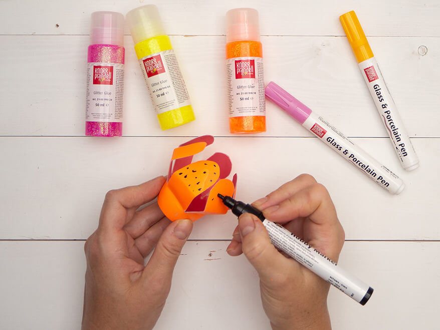 Jemand verziert eine, zu einer orangenen Blume upcycelte Plastikflasche mit einem Acrylmarker. Drumherum liegen weitere Marker und verschiedene Glitzerkleber von Knorr Prandell. Der Untergrund ist hell. Foto: Johanna Rundel
