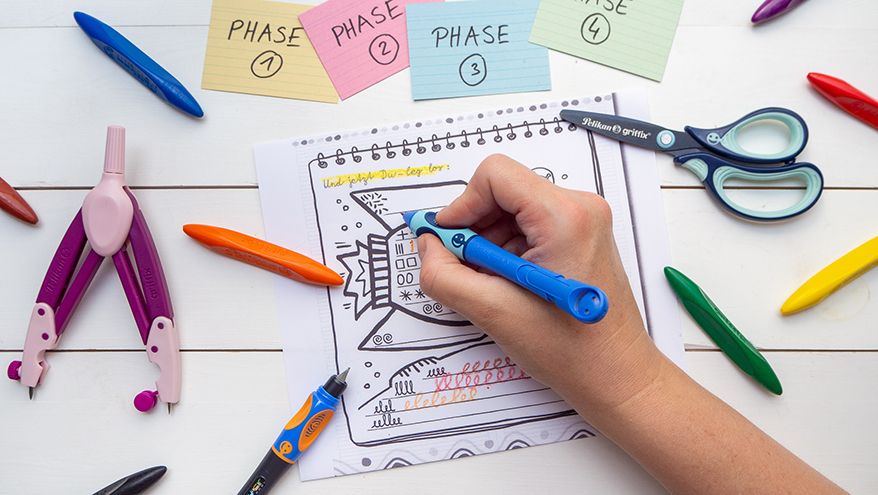 Jemand schreibt mit einem Schreiblernstift von Pelikan auf ein Blatt mit Schwungübungen. Drumherum liegen ein lila Zirkel, eine blaue Schere, mehrere verschiedenfarbige, dreieckige Wachsmalkreiden und bunte Karteikarten. Der Untergrund besteht aus weißem Holz. Foto Johanna Rundel 