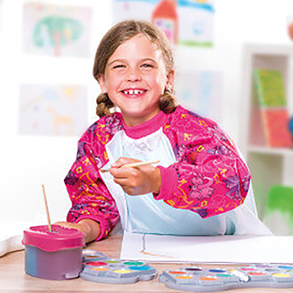 Ein Mädchen im Vorschulalter malt mit Wasserfarben auf ein weißes Blatt Papier. Es trägt dabei eine pinke Malschürze und lacht in die Kamera. Im Hintergrund sind selbstgemalte Bilder zu sehen.