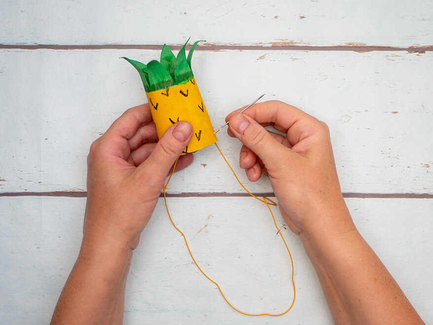Jemand fädelt eine dünne, gelbe Gummikordel in eine zur Ananas umgestalteten, leeren Klopapierrolle. Es wird ein Partyhut. Der Untergrund ist hell. Foto: Johanna Rundel