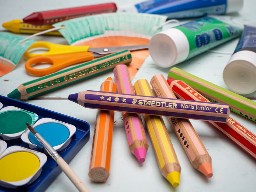 Auf einem hellen Untergrund liegen verschiedene Bastelmaterialien von Staedtler: dicke Buntstifte, Wachsmalstifte, Schere, ein Wasserfarbkasten, Fingerfarben und ein Pinsel. Foto: Johanna Rundel 