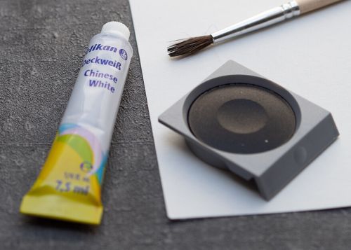 Auf einem dunkelgrauen Untergrund liegt ein Blatt Papier. Darauf liegt ein Pinsel, eine Tube Deckweiß und ein Näpfchen schwarze Deckfarbe von Pelikan. Foto: Johanna Rundel