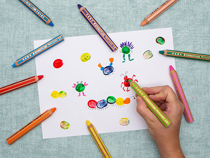 Jemand malt mit einem hellgrünen, dicken Buntstift von Staedtler Details an einen roten Farbfleck (ein Fingerabdruck aus Fingermalfarbe) auf einem weißen Blatt Papier. Drumherum sind weitere, in dieser Art, gestaltete "Tierchen". Auf dem türkisfarbenen Untergrund liegen weitere Buntstifte in verschiedenen Farben. Foto: Johanna Rundel