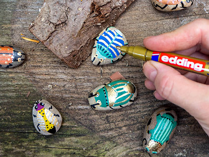 Auf einem rustikalen Holzuntergrund liegen mehrere Steine, die als Käfer bemalt wurden. Von rechts ragt eine Hand ins Bild, die mit einem Metallicmarker der Marke edding einen der Steine bemalt. Foto: Johanna Rundel