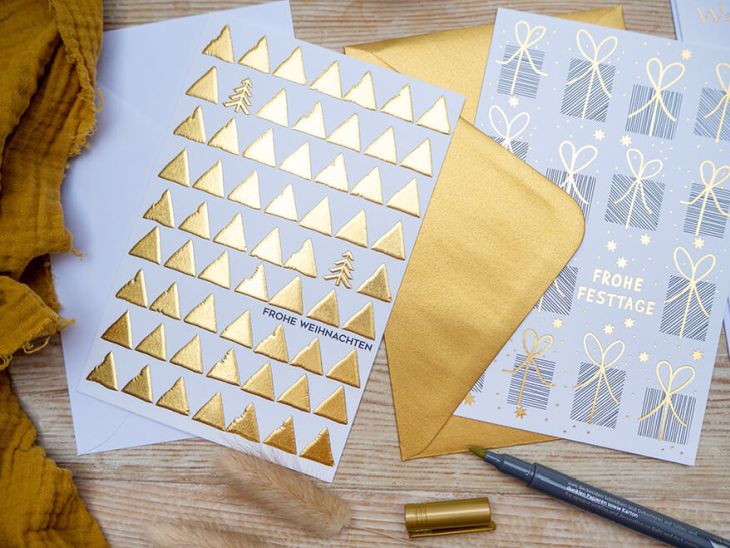 alt="Auf einem Holztisch liegen verschiedene Weihnachtkarten von ABC Grußkarten, sowie ein goldener Briefumschlag. Die Karten sind mit aufwändiger Goldprägung in verschiedenen Designs gestaltet. Ein geöffneter goldener Brushpen liegt daneben. Foto: Johanna Rundel"
