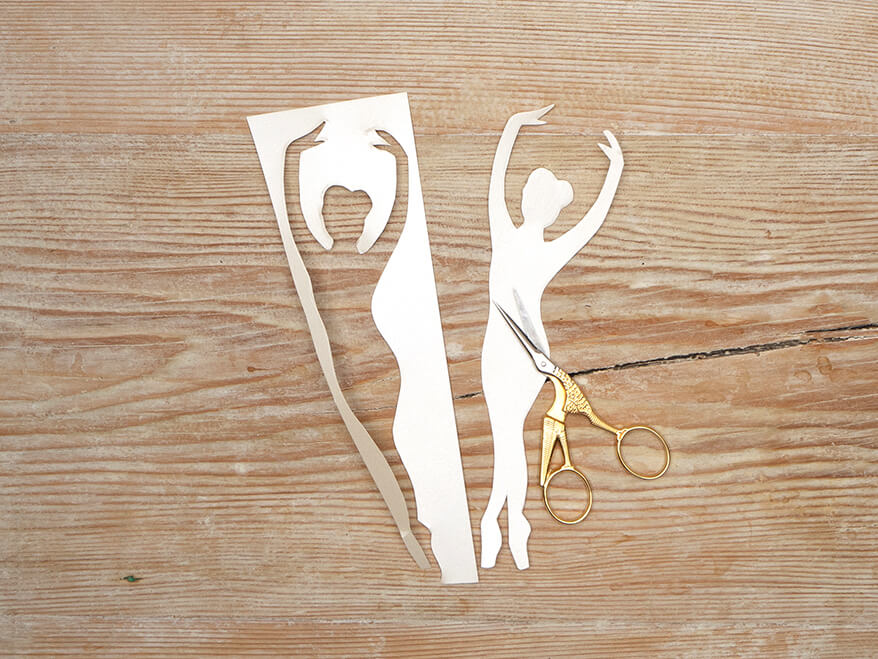 Aus einem hellen Papier von Artoz wurde die Silhouette einer Tänzerin ausgeschnitten. Die goldene Kranichschere liegt auf der Figur. Die Papierreste daneben. Der Untergrund ist ein rustikales, helles Holzbrett. Foto: Johanna Rundel