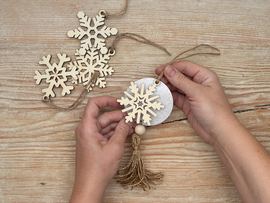 Jemand bastelt aus einem Papierkreis, auf dem eine Holzschneeflocke liegt und einer goldenen Tassel einen Geschenkanhänger. Daneben liegen weitere Holzschneeflocken von Artoz. Der Untergrund ist ein rustikales, helles Holzbrett. Foto: Johanna Rundel