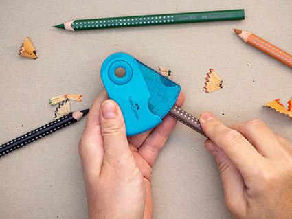 Jemand spitzt dicke Buntstifte von Faber-Castell in einem türkisfarbenen Klappspitzer an. Drumherum liegen die Spitzer-Reste. Der Untergrund ist ein graubraunes Kraftpapier. Foto: Johanna Rundel