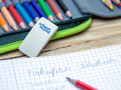 Auf einem Holztisch liegt ein aufgeschlagener, karierter Collegeblock. Darauf wurde mit einem roten Bleistift eine Einkaufliste geschrieben. Dahinter sieht man ein aufgeschlagenes, grünes Schülermäppchen mit Buntstiften und einen Radiergummi von School-Mood. Foto: Johanna Rundel