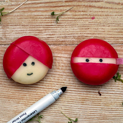 Auf einem Holzbrett liegen zwei Mini-Käse mit roter Wachshülle. Die Hülle wurde so geformt, dass aus einem Käse ein Gesicht mit Turban und aus dem anderen ein Ninja geworden ist. Mit einem schwarzen Lebensmittelstift wurde den runden Käseköpfen Augen gemalt. Foto: Johanna Rundel