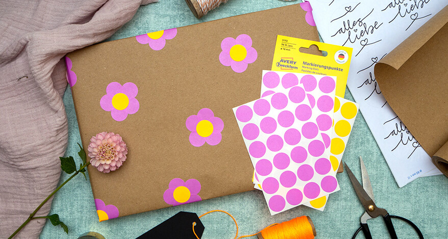 Auf einem türkisfarbenen Untergrund liegt ein in Kraftpapier eingepacktes Geschenk. Darauf sind mit pinken und neonorangen Markierungspunkten von Avery Zweckform kleine Blumen geklebt. Auf dem Geschenk liegen weitere Aufkleberbögen mit Markierungspunkten und eine rosafarbene Dahlienblüte. Unter dem Geschenk liegt eine Rolle neonoranges Bäckergarn, eine schwarze Schere und ein schwarzes Tafeletikett. In der linken, oberen Ecke wurde rosa Stoff drapiert. In der rechten liegen runde Aufkleberbögen und eine Rolle Kraftpapier. Foto: Johanna Rundel
