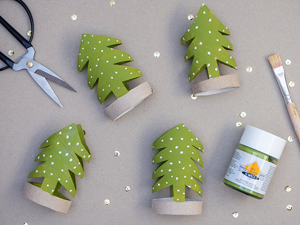 Auf einem grauen Untergrund liegen vier grüne Tannenbäume, die aus Klopapierrollen geschnitten wurden. Eine schwarze Schere, ein Malpinsel und ein Gläschen grüne Farbe von Baier und Schneider liegen daneben. Goldene Pailletten sind über den gesamten Untergrund verteilt. Foto: Johanna Rundel