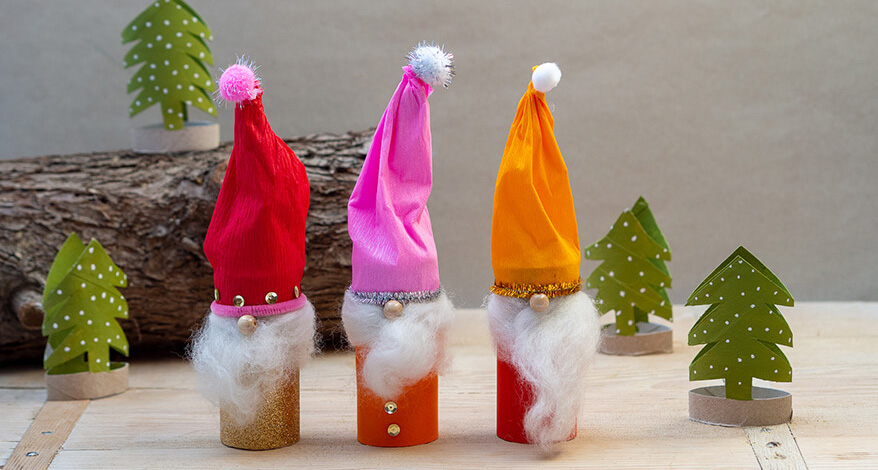 Vor einem grauen Hintergrund stehen drei Weihnachtswichtel, die aus Klopapierrollen und Materialien von Baier und Schneider gebastelt wurden. Außerdem sind vier grüne Tannenbäum, die aus Klopapierrollen geschnitten wurden, zu sehen. Im Hintergrund liegt ein Holzstamm. Der Untergrund ist ein rustikales Holzbrett. Foto: Johanna Rundel