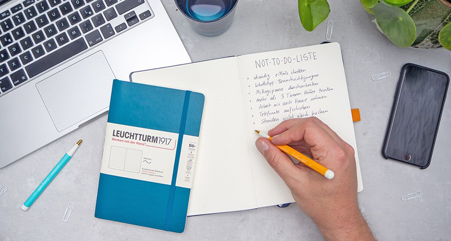 Jemand schreibt mit einem orangenen Drehgriffel eine Not-To-Do-Liste in ein Bullet Journal von Leuchtturm1917. Auf dem aufgeschlagenen Journal liegt ein petrolfarbenes Notizbuch. Daneben ist ein weiterer Drehgriffel in türkis, ein aufgeklappter Laptop, ein Glas Wasser, eine Grünpflanze und ein schwarzes Smartphone zu sehen. Einige weiße Büroklammern liegen verstreut darum. Der Untergrund ist hellgrau. Foto: Johanna Rundel 