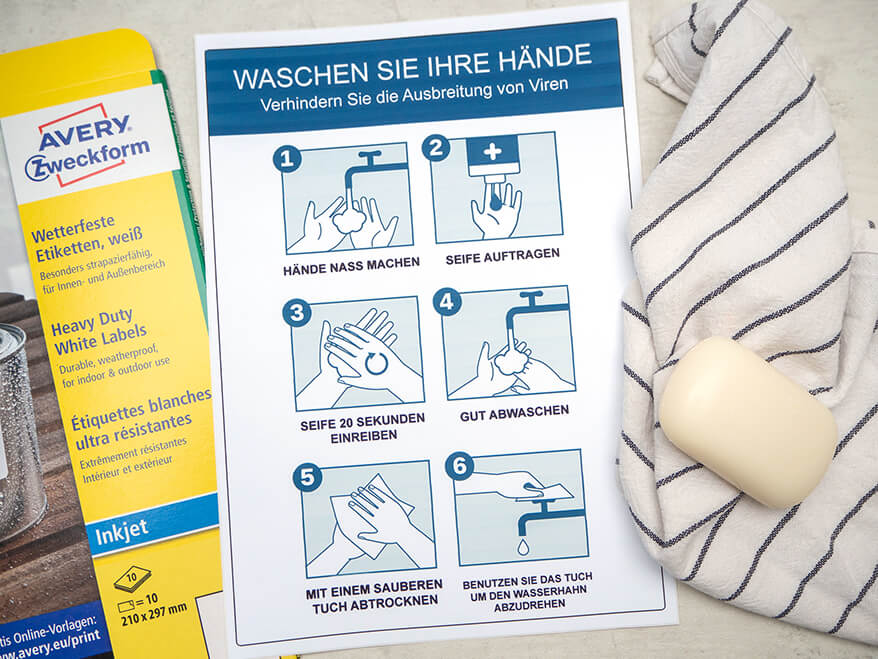 Auf einem grauen Untergrund in Betonoptik liegt eine Packung weiße, wetterfeste Etiketten von Avery-Zweckform. In der Mitte ist ein Hinweisschild zum Händewaschen zu sehen, rechts davon ein Handtuch, auf dem ein Stück Seife liegt. Foto: Johanna Rundel