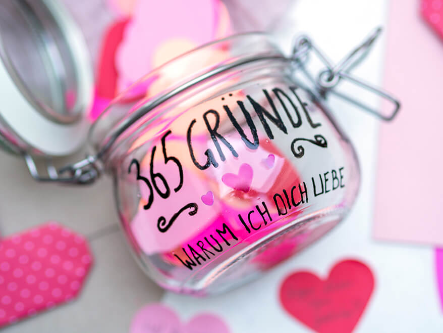 Auf einem hellen Untergrund liegt ein geöffnetes Vorratsglas, auf das mit einem schwarzen Glass & Porcelain Pen von Knorr Prandell "365 Gründe, warum ich dich liebe" geschrieben wurde. Aus dem Glas fallen viele Papierherzen in Rosa- und Rottönen. Foto: Johanna Rundel