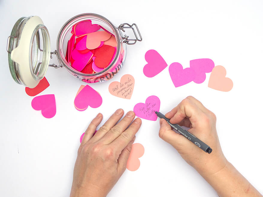 Mit einem Stift werden Botschaften auf Papierherzen in Rosa- und Rottönen geschrieben. Einige Herzen sind in einem Vorratsglas gesammelt. Foto: Johanna Rundel