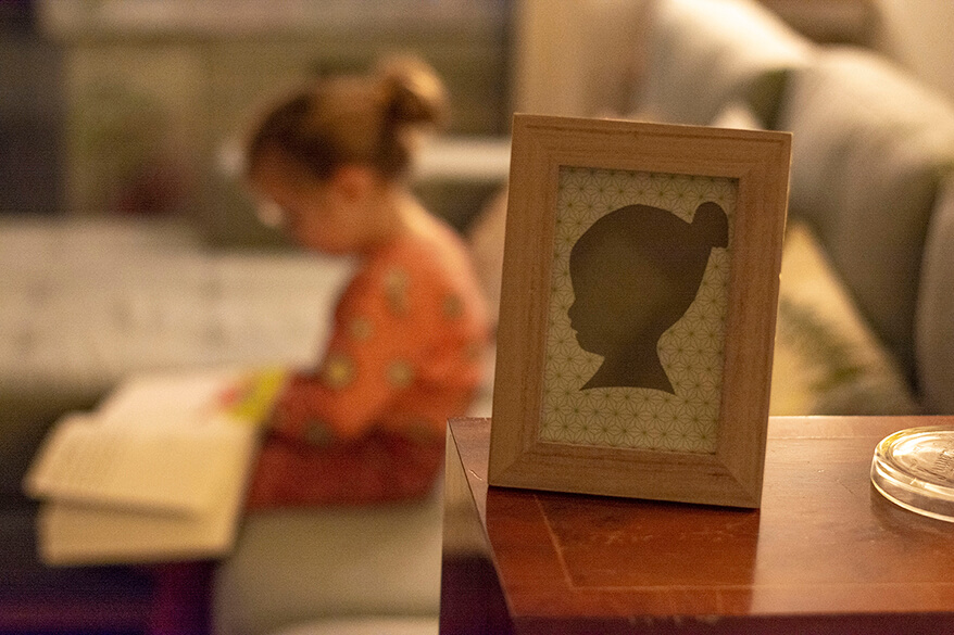 Im Vordergrund steht ein Do-it-yourself-Scherenschnitt dem Kopf eines Mädchens in einem Bilderrahmen auf einem Tisch. Im Hintergrund sieht mal verschwommen das gleiche Mädchen in echt auf einem Sofa sitzen. 