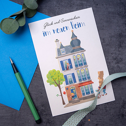 Auf einem anthrazitfarbenen Untergrund liegt eine Grußkarte des ABC Glückwunschkartenverlags, daneben ein blauer Briefumschlag und ein grüner Stift. Auf der Karte ist ein hohes, buntes Stadthaus zu sehen. Zwei Personen schleppen Umzugskarton hinein. Auf der Karte steht: "Glück und Sonnenschein im neuen Heim". Foto: Johanna Rundel