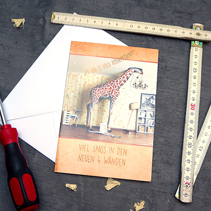 Auf einem anthrazitfarbenen Untergrund liegt eine Grußkarte des ABC Glückwunschkartenverlags auf einem weißen Briefumschlag. Auf der Karte ist eine Giraffe zu sehen. Der Spruch "Viel Spaß in den neuen vier Wänden" ist darauf gedruckt. Neben der Karte liegen ein ausgeklappter Zollstock und ein roter Schraubendreher, sowie einzelne Holzspäne. Foto: Johanna Rundel