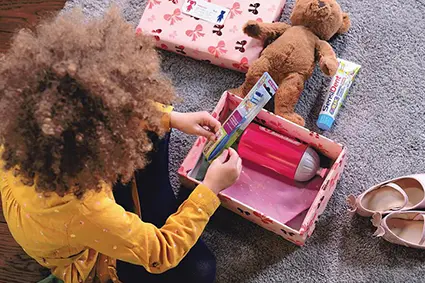Ein Kind mit wilden, dunklen Locken und gelben Oberteil packt eine Geschenkschachtel aus. Darin liegt eine pinke Trinkflasche aus Plastik von Step by Step. Drumherum liegen weitere Geschenke aus der Schachtel. Das Kind hält eine Zahnbürste in der Hand.