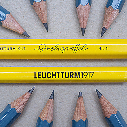 Auf einem grauen Untergrund liegen horizontal ein gelber Drehgriffel und ein gelber Bleistift von Leuchtturm1917. Acht blaue Bleistifte liegen im Kreis darum. Ihre Spitzen zeigen auf die Schriftzüge der gelben Stifte. Foto: Johanna Rundel