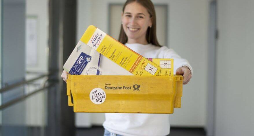 Eine Frau hält gelbe Postkiste mit Etiketten und Briefumschlägen in die Kamera.