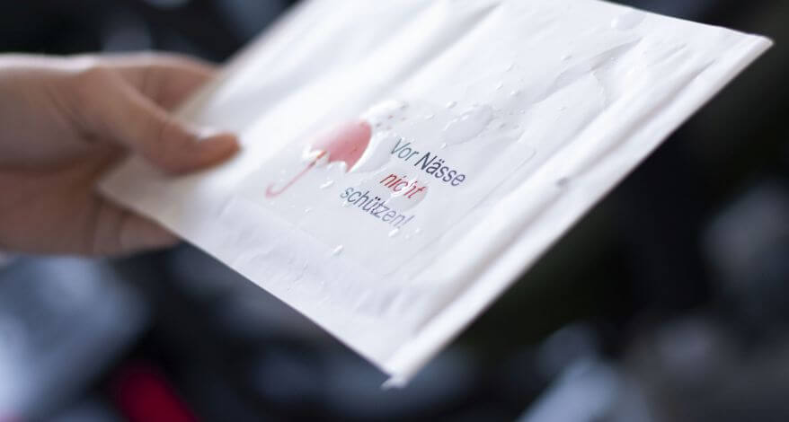 Nahaufnahme eines wetterfesten Etiketts von Avery Zweckform, das auf eine weiße Versandtasche geklebt ist. Eine Hand hält den Umschlag fest, auf dem Etikett sind Wassertropfen zu sehen, die das Licht reflektieren. Auf dem Etikett ist ein roter Regenschirm abgedruckt und der Text „Vor Nässe nicht schützen“.