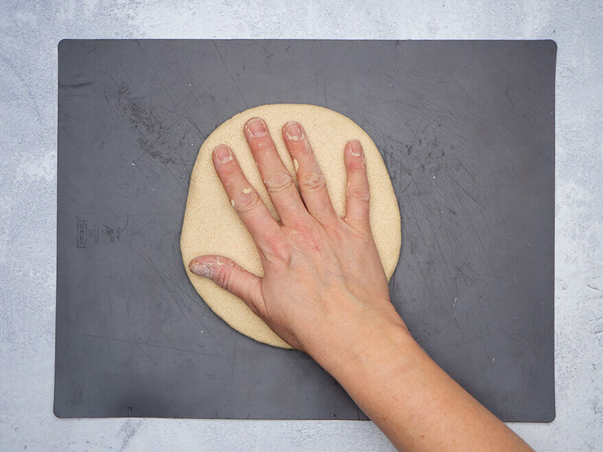Jemand drückt seine Handfläche in einen Kreis aus Salzteig, der auf einer dunkelgrauen Backmatte liegt. Der Untergrund ist Betongrau. Foto: Johanna Rundel 