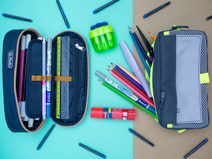 Zwei Faulenzermäppchen von Herlitz liegen nebeneinander. Aus dem rechten, in Grautönen mit neongrünen Akzenten, zeigen Buntstifte, Schere, Tintenlöscher und ein Füller von Pelikan heraus. Das linke, in dunkelblau mit einem braunen Reißverschluss, ist ebenfalls mit Stiften und Schulmaterialien gefüllt. Auf dem türkis-grauen Untergrund liegen in unregelmäßigen Abständen lange, blaue Tintenpatronen. Foto: Johanna Rundel