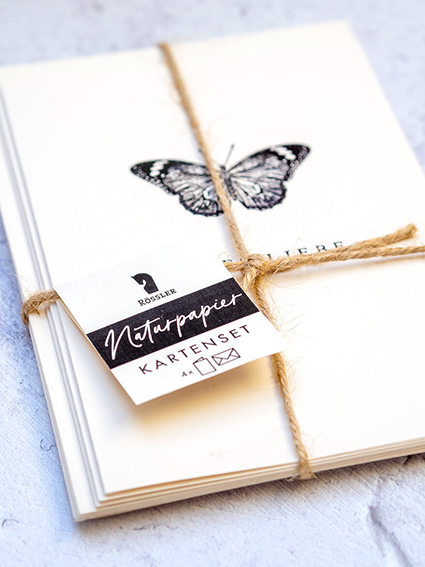 Auf einem hellgrauen Untergrund liegt ein mit Jutefaden zusammengebundenes Bündel Grußkarten aus Naturpapier von Rössler. Darauf ist ein schwarzer Schmetterling zu sehen. Foto: Johanna Rundel