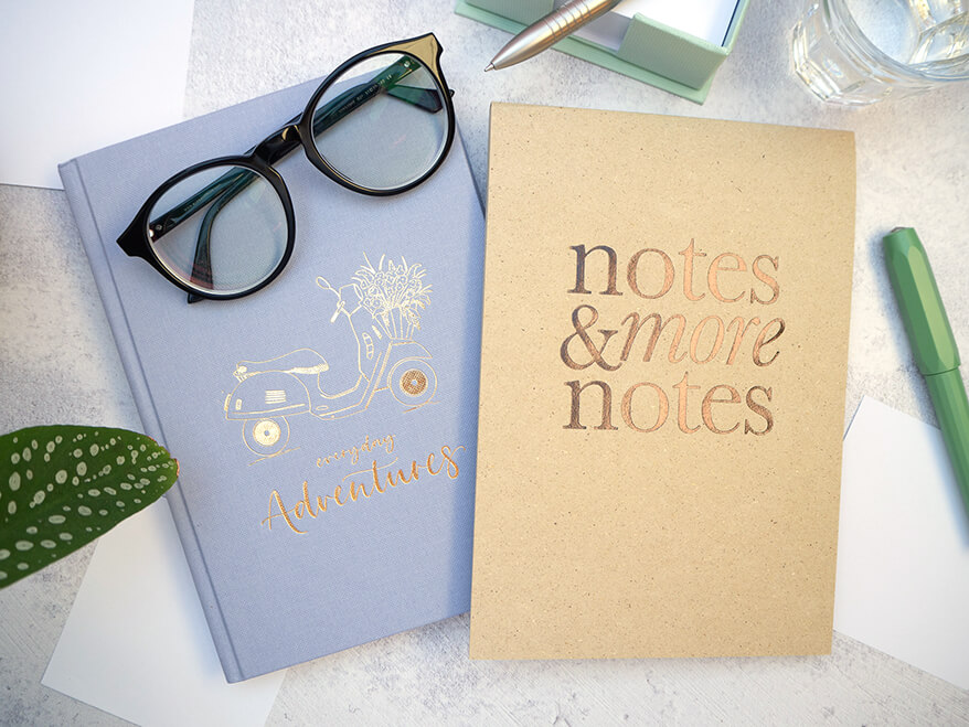 Auf einem hellgrauen Untergrund liegen ein hellblaues Journal und ein Notizblock aus Kraftpapier von Rössler. Auf dem Journal ist eine Vespa abgebildet. Auf dem Block steht in goldener Schrift „notes & more notes“. Auf dem Journal liegt eine schwarze Brille. Drumherum sind mehrere weiße Notizzettel verteilt. Der dazugehörige mintfarbene Zettelkasten ist darüber zu sehen. Foto: Johanna Rundel