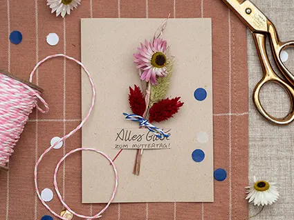 Auf einer altrosa Tischdecke mit weißen Streifen, liegt eine selbstgebastelte Muttertags Grußkarte aus Kraftpapier. Auf der Karte steht in schwarzem Lettering "Alles Gute zum Muttertag". Ein Sträußchen Trockenblumen von Artoz ist in einen Schlitz in der Karte gesteckt worden. Um die Karte herum liegt Konfetti und weitere Trockenblumen. Außerdem ist eine goldene Schere, sowie eine Rolle rosa-weiß gestreiftes Bäckergarn im Bild zu sehen. Foto: Johanna Rundel