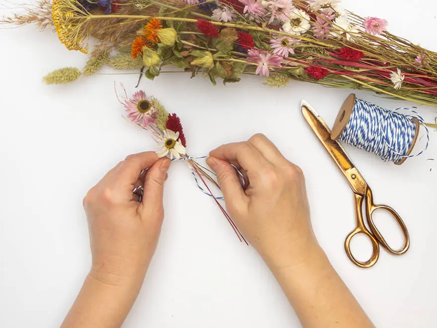 Jemand bindet ein kleines Sträußchen aus Trockenblumen von Artoz. Es wird mit blau-weiß gestreiftem Bäckergarn zusammengebunden. Das Garn, sowie eine goldene Schere, sind zusammen mit dem großen Blumenbündel ebenfalls im Bild zu sehen. Der Untergrund ist weiß. Foto: Johanna Rundel