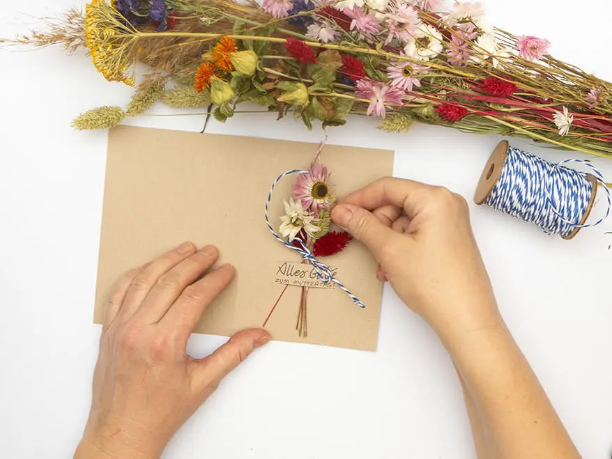 Jemand steckt ein kleines Sträußchen Trockenblumen in einen Schlitz in einer Klappkarte aus Kraftkarton von Artoz. Der Untergrund ist weiß. Foto: Johanna Rundel