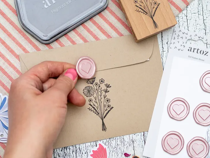 Jemand klebt einen Wachssiegel-Aufkleber auf einen Briefumschlag aus Kraftpapier von Artoz. Der Bogen mit weiteren Aufklebern liegt rechts daneben. Oberhalb des Umschlags ist ein schwarzes Stempelkissen und ein Holzstempel mit Blumenmotiv zu erkennen. Der Untergrund ist ein rosa-beige gestreiftes Tischtuch. Foto: Johanna Rundel