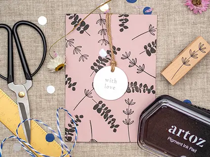 Auf einem Juteuntergrund liegt eine altrosafarbene Klappkarte von Artoz, die mit botanischen Motiven (Eukalyptuszweige und Gräser) bestempelt wurde und auf die ein weißer Geschenkanhänger mit der Aufschrift „with Love“ befestigt wurde. Drumherum sind verschiedene Utensilien, die zu Herstellung der DIY Muttertagskarten verwendet wurden, verteilt: schwarzes Stempelkissen, Stempel, Schere und ein goldenes Lineal. Foto: Johanna Rundel