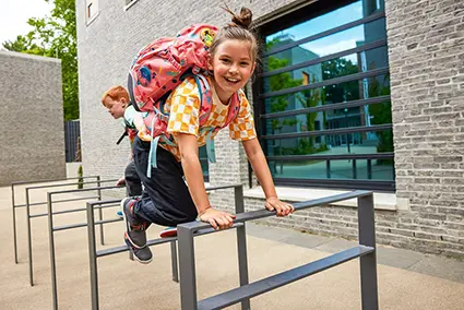 Ein Mädchen im Grundschulalter springt über einen anthrazitfarbenen Fahrradständer vor einem grauen Schulgebäude. Im Hintergrund ist ein rothaariger Junge zu sehen, der ebenfalls auf den Fahrradständern herumklettert. Beide tragen Schulranzen von ergobag auf dem Rücken.
