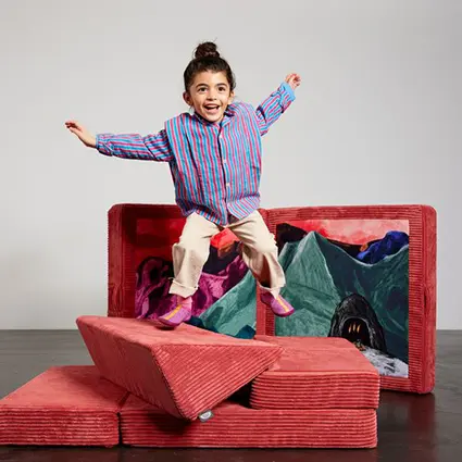 Ein Mädchen in einem rot blau gestreiften Hemd hüpft fröhlich auf einem roten Kindersofa aus Cord von Affenzahn herum.