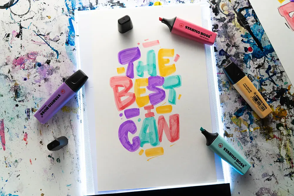 Auf einem Leuchttisch liegt ein DIN A4 Blatt mit einem bunten Buchstaben-Lettering. Drumherum liegen verschiedene Marker von Stabilo.